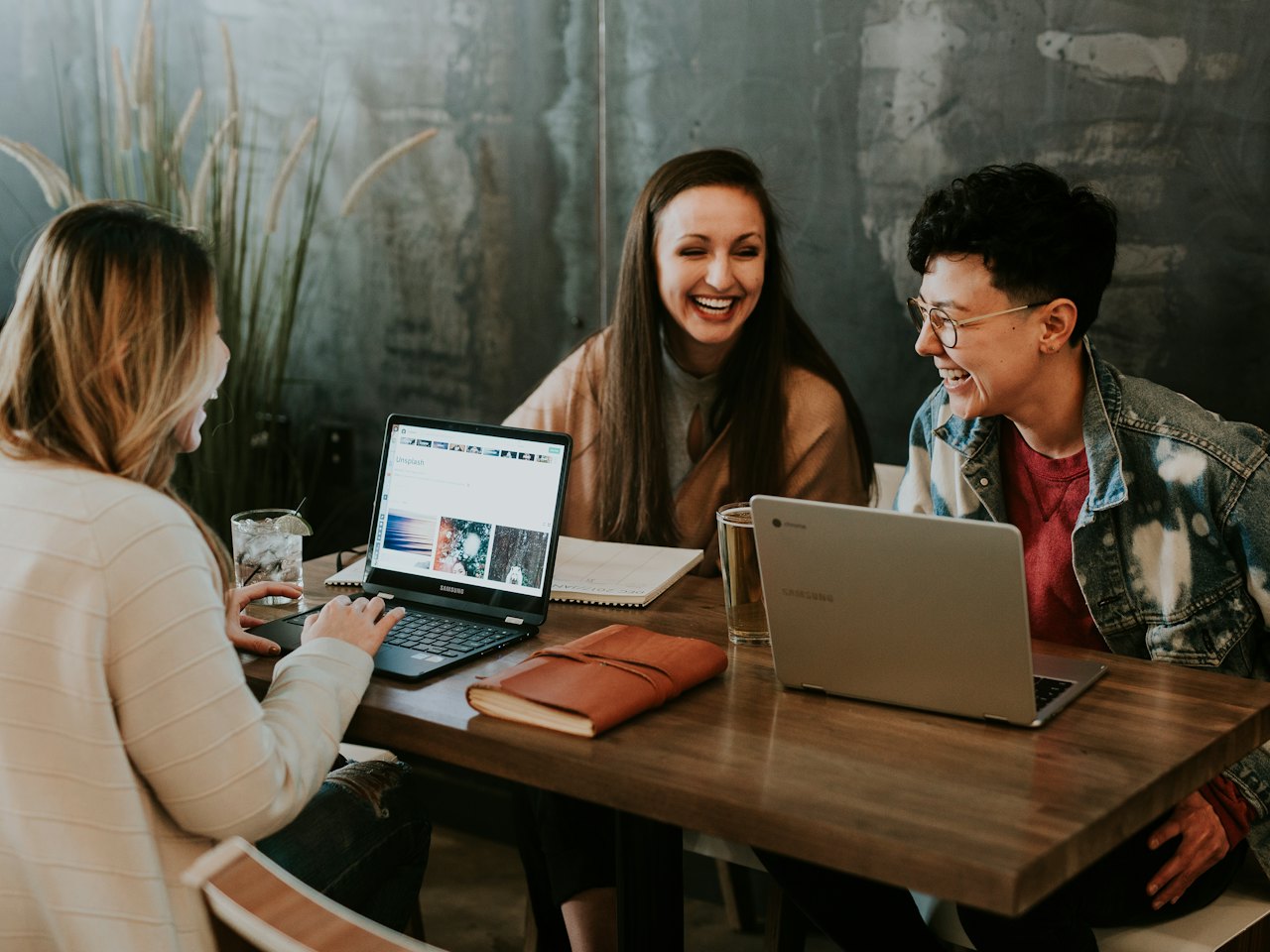 Three adult learners talking and smiling around a laptop at a café table