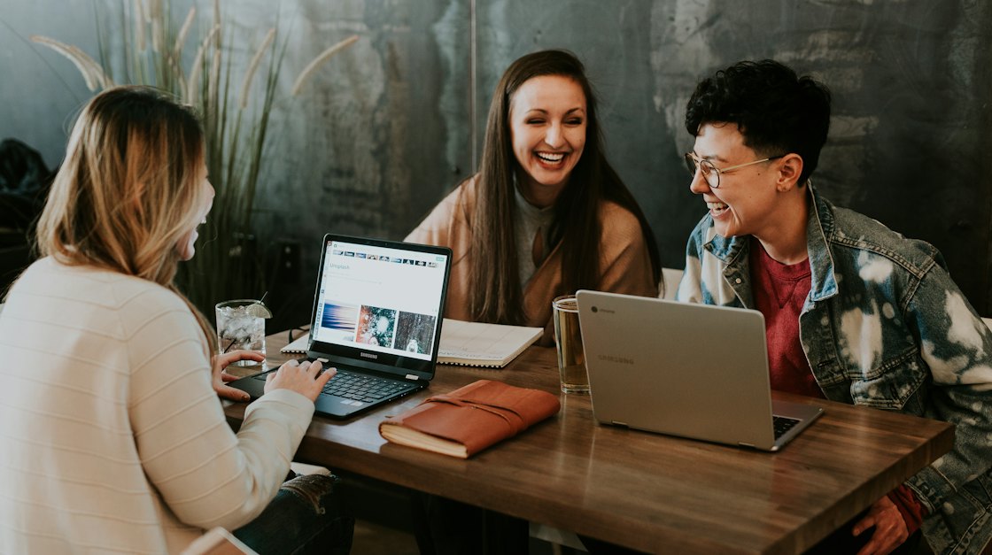 Three adult learners talking and smiling around a laptop at a café table